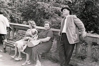 A man in a tweed suit and hat observes two women seated on a bench. The women are dressed in 1950s style attire and one holds...