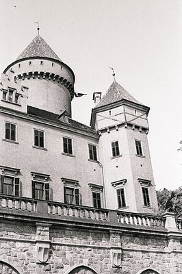 Exterior view of a castle with a central round tower and a square tower with a steeply pitched roof, both topped with decorat...