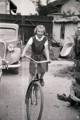 Young girl on a vintage bicycle, captured in motion. Classic 1950s car visible in the background along with simple housing/ou...