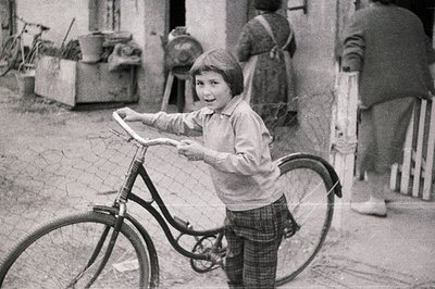 A young boy smiles while holding a vintage bicycle, standing outside a rural home. The home's exterior shows weathered siding...