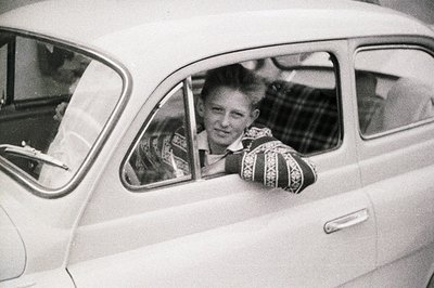 Young man leans out of a vintage Volkswagen Beetle, likely 1960s, exhibiting a casual, relaxed pose. Patterned sweater & shor...