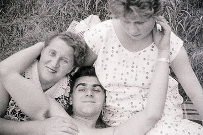 A black and white candid photo depicts a man lying on grass embraced by two women. All three appear relaxed and smiling. The ...