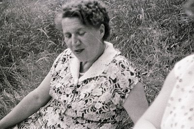 Close-up, black and white image of a woman seated in tall grass. She wears a short-sleeved, patterned dress with a Peter Pan ...