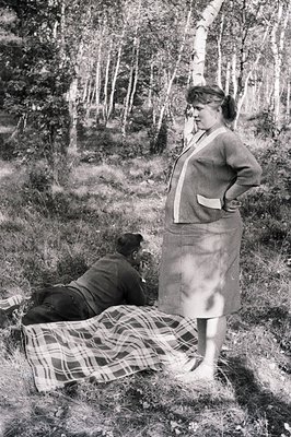A man reclines on a checkered blanket in a birch forest, absorbed in reading. A woman stands nearby, arms crossed, seemingly ...