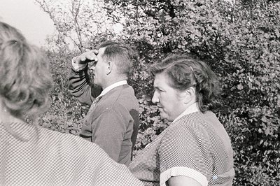 A man in a tweed jacket and collared shirt, peering through what appears to be a telescope, stands next to a woman with a pat...