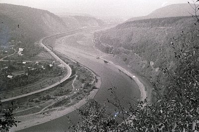 Monochrome aerial view depicts a winding river valley, likely a substantial river with barges visible. A curving roadway para...
