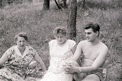 A candid snapshot captures three individuals seated on a blanket amidst tall grass and a blurred treeline. The group appears ...