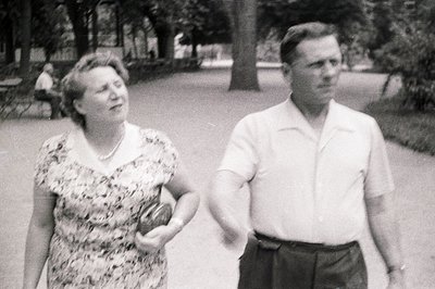 A couple walks along a tree-lined path in a park. The woman wears a patterned dress & pearl necklace; the man, a short-sleeve...