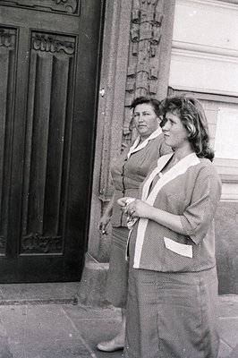 Two women stand outside a building with an ornate door. Both are dressed in matching striped suits with wide collars. Appears...