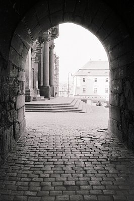 View through an arched stone passageway reveals a public square with tiered steps, a columned portico, and a building with si...