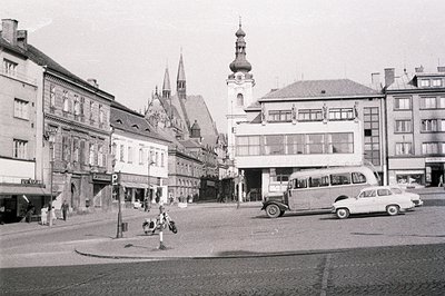B&W photo depicts a European town square, showcasing a mix of architectural styles. Visible are ornate facades, a church stee...