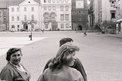 Formal architectural facade dominates a cobblestone square. Three figures are in the foreground, partially obscuring the view...