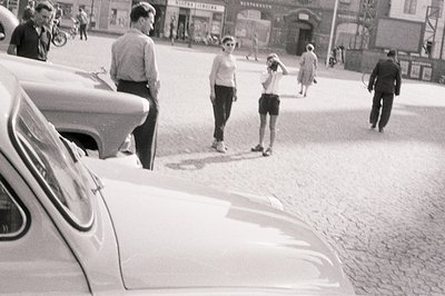 A monochrome street scene captures a group of people in what appears to be a European city. A classic car, possibly a Fiat, d...
