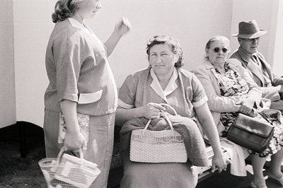 A group gathers, possibly awaiting an event or service. A woman in a striped suit gestures, while others sit with woven bags ...