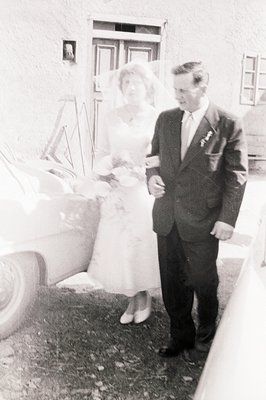 A bride and groom stand beside a classic car, likely post-ceremony. The woman wears a full-skirted wedding dress and veil; th...