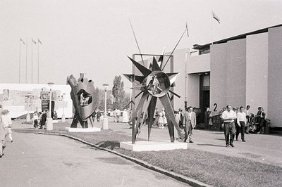 Monochrome view of a 1960s exhibition space. Abstract sculptures dominate the foreground, with a modernist pavilion visible t...
