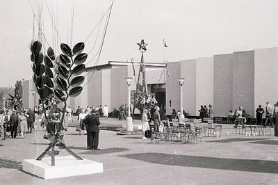 Monumental sculpture of stylized foliage on a plinth dominates this view of a public space, likely an exposition or fair. A s...