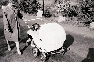 A woman in a long coat pushes a distinctive, rounded baby carriage along a rural road. The carriage features a textured canop...