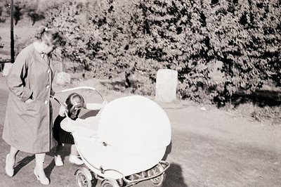 A woman in a knee-length coat pushes a unique, futuristic-looking baby carriage featuring a round basin and chrome accents. T...