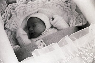 Infant sleeps peacefully in a lace-trimmed cot, swaddled in white cloth. The photograph's grain suggests a vintage print, lik...