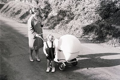 Vintage photograph showing a woman and child in a distinctive wicker baby carriage. The woman wears a long coat & heels; the ...