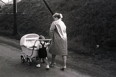 A woman in a trench coat walks along a rural road, pushing an ornate wicker baby carriage. The child, dressed in a dark dress...