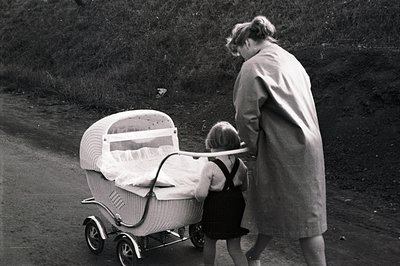 A rear view captures a woman in a long coat pushing a distinctive, woven wicker baby carriage along a gravel road. A child st...