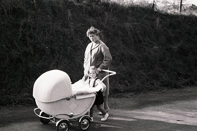 A woman in a tailored suit pushes a vintage wicker baby carriage, with a young child visible within. The scene takes place on...