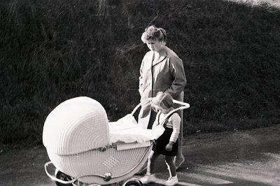 A woman in a long coat pushes a distinctive wicker baby carriage along a paved road. The carriage's curved design and ornate ...