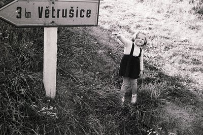 A young girl in a dark dress and bloomers poses next to a weathered signpost indicating "3 km Větrusice." The monochrome imag...