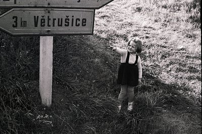 Young girl in a dark dress and tights stands beside a signpost indicating "Větrušice, 3km". The photo, likely from the 1960s ...