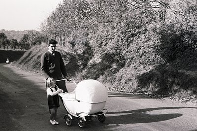 A young man in a suit and tie guides a child with a unique, rounded baby carriage along a tree-lined road. The carriage's des...