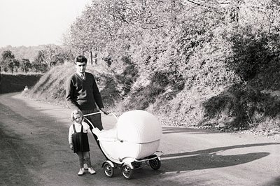 Man in a sweater and tie stands with a young girl beside an unusual, futuristic-looking pram on a rural road. Likely mid-20th...