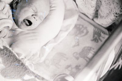 A baby in a crib, captured in a candid moment of distress or surprise. The child's mouth is open in a cry or gasp. Crib raili...