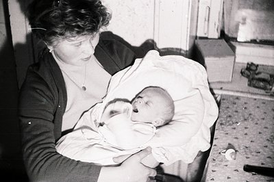 A woman holds a swaddled infant in her arms, viewed from above. Interior setting; likely a domestic space with a tiled floor ...