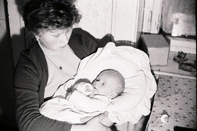 A young woman holds a swaddled infant close, viewed in a dimly-lit, interior setting. She wears a dark cardigan and a simple ...