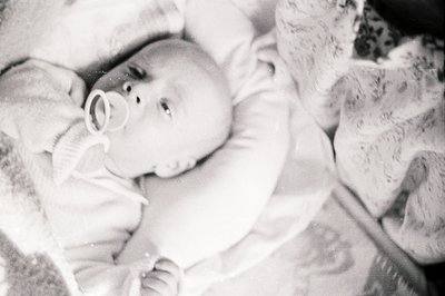 Close-up black and white photograph of an infant, likely a baby, bundled in a textured blanket. The child holds a pacifier in...
