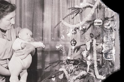 A mother gently holds a baby in front of a decorated Christmas tree. The tree features tinsel, glass ornaments, and tradition...