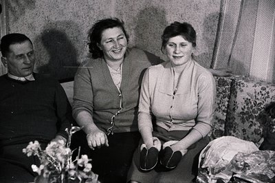 A black and white snapshot of a man, woman, and young woman seated on a floral patterned sofa. The man wears a dark sweater. ...