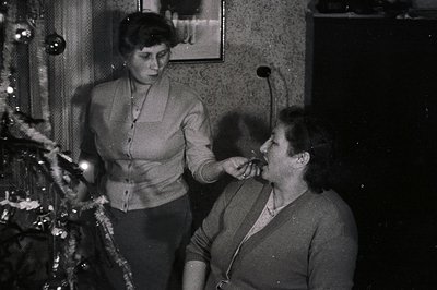 Two women near a decorated Christmas tree. One offers the other a cigarette. Interior scene with textured wallpaper & traditi...