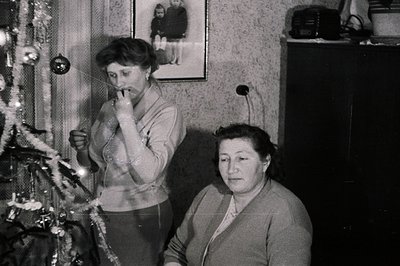 Black and white photo depicting two women indoors, one decorating a Christmas tree, the other seated, seemingly observing. Mi...