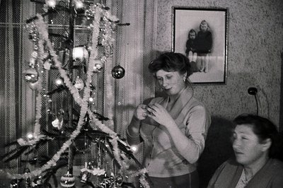 A woman decorates a sparsely-ornamented Christmas tree, possibly in the 1950s or 60s. A framed portrait of two children hangs...