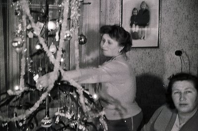 A woman decorates a Christmas tree with garlands, captured in a candid, snapshot-style image. A second woman observes from th...