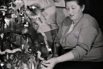A woman decorates a Christmas tree with string lights, captured in a candid moment. The tree features glass ornaments. Interi...