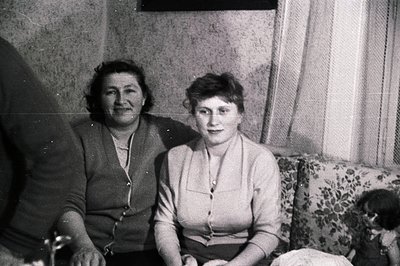 Two women seated on a floral patterned sofa. The woman on the left wears a cardigan over a high-necked blouse, while the woma...
