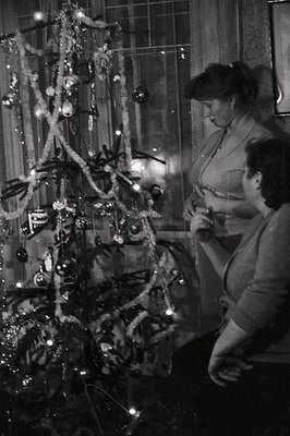 Two women decorate a tinsel-laden Christmas tree indoors, likely in the 1950s or 60s. Natural light and decorative lights ill...