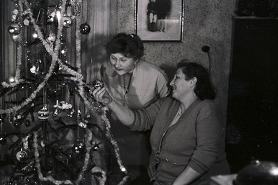 Two women decorate a Christmas tree in a cozy, modestly furnished room. A framed portrait hangs above the tree, suggesting a ...