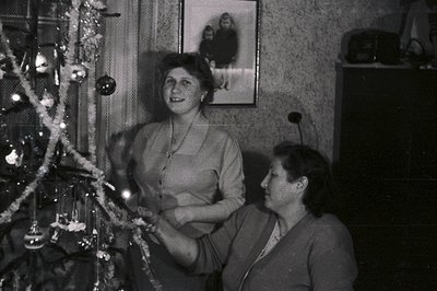 Two women decorate a Christmas tree in a modestly furnished room. A framed portrait hangs on the wall. Likely a 1950s or 60s ...