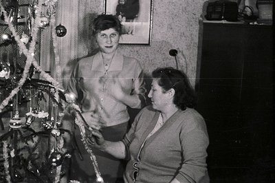 A black and white snapshot captures two women decorating a Christmas tree indoors. One stands, adjusting a garland; the other...