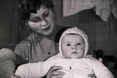 A woman cradles a baby bundled in a heavily textured, knitted bonnet and sweater. Black and white photography suggests a mid-...